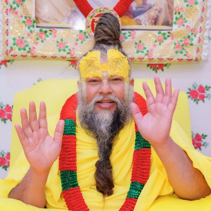 Premanand Ji Maharaj seated on his asan with both hands raised, giving darshan to devotees at satsang