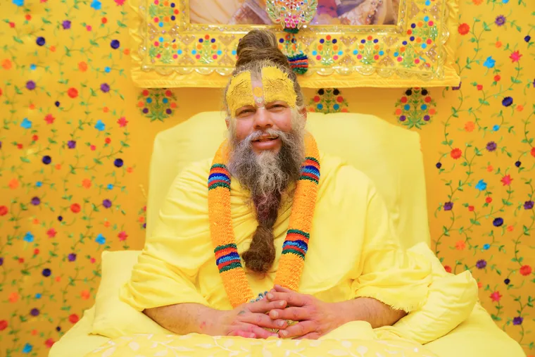 Premanand Ji Maharaj seated on his asan in yellow robes before a decorated backdrop at Shri Hit Radha Keli Kunj, Vrindavan