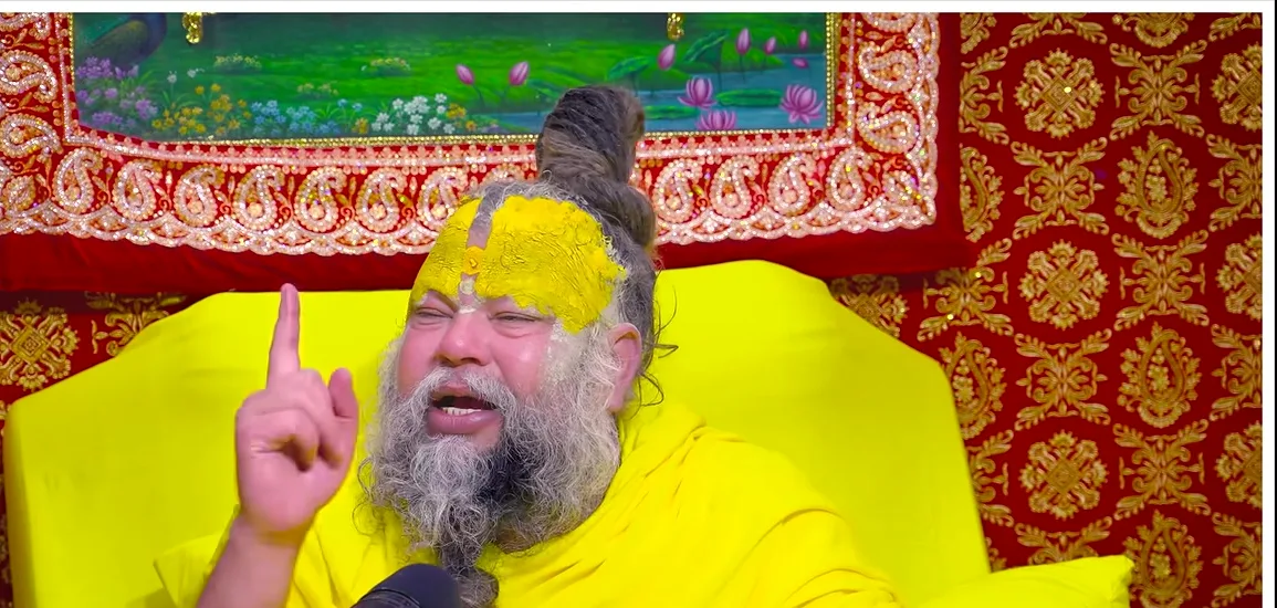 Premanand Ji Maharaj one finger raised before a red backdrop