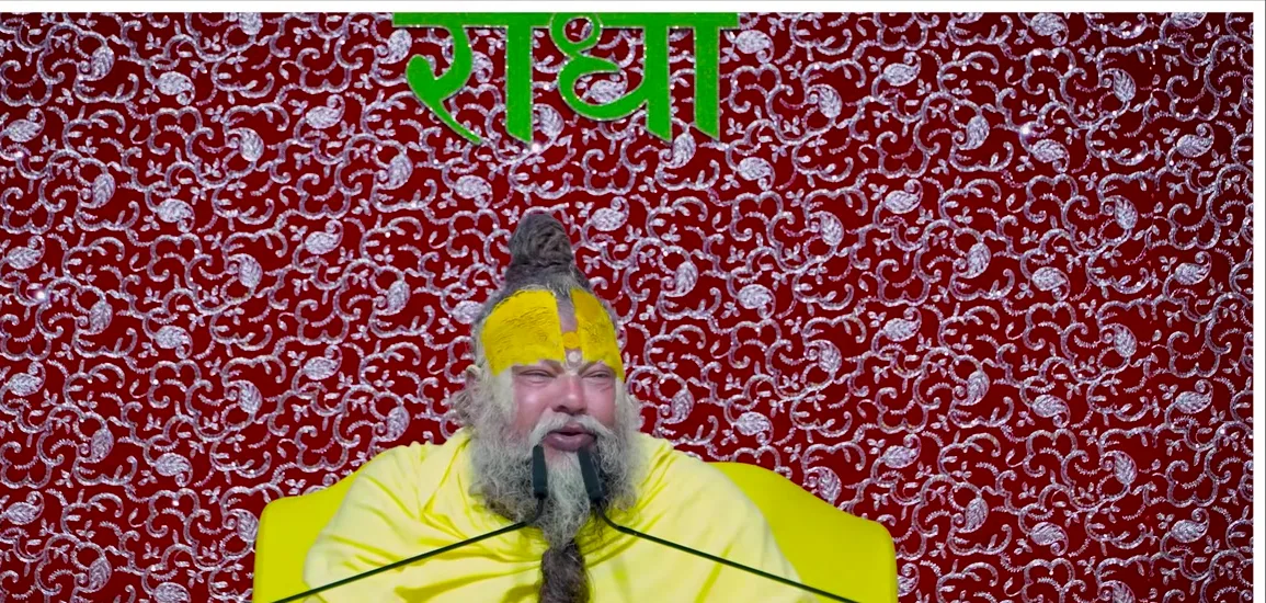 Premanand Ji Maharaj seated before a deep red backdrop bearing the name Radha