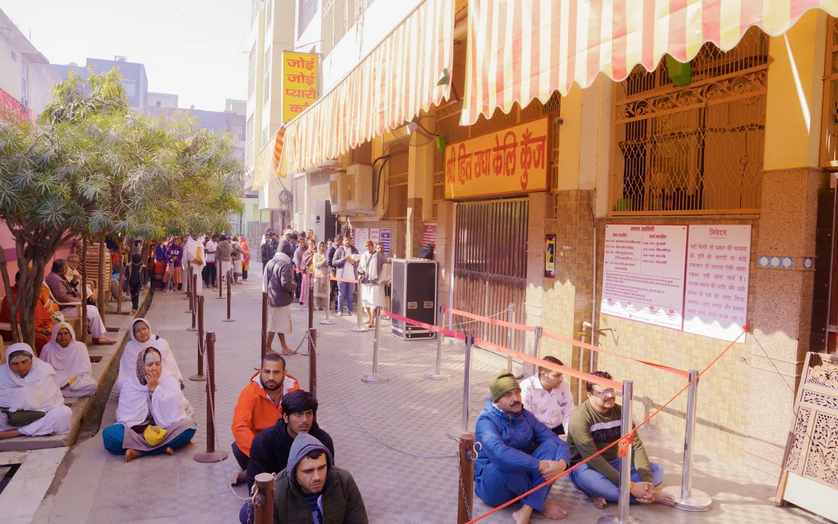 Devotees seated in queue along the lane outside Shri Hit Radha Keli Kunj ashram in Vrindavan, with the timing board on the wall